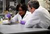 An image showing Malika Jeffries-EL with 'Beyond the Elements' presenter David Pogue. Both are wearing white lab coats. Malika is holding long tweezers with which she is pulling a stringy substance - nylon - out of a beaker filled with a reddish liquid.