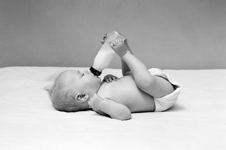 A black and white image showing a baby lying on his back and feeding himself from a bottle held by his legs