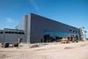 A photo of a grey blocky building with a large glass front under a bright blue skye. The building is surrounded by construction sand, there is a digger standing in front of the building and the top of a crane is poking over the top of the roof.