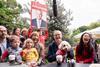 A white, middle-aged, bespectacled man seated at a table. He's laughing and holding a dog on his lap. There is a small crowd of people gathered to his right, one holding an election campaign poster.