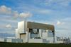 A photo of a white large blocky building, surrounded by smaller blocky buildings set behind a barbed wire fence. It's a clear sunny day and there are fluffy clouds in the sky.