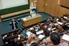 A man stands at the front of a lecture theatre in front of several rows of students sat with papers on the benches in front of them. On the blackboard behind him is a chemical structure