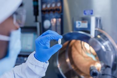 A gloved hand holds a pill up to the camera in a pharmaceutical lab