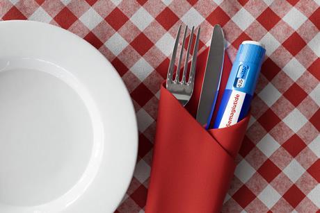 A semaglutide injecting 'pen' wrapped in a napkin with a knife and fork, on a red gingham tablecloth, next to a white plate