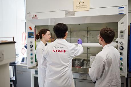 A member of staff at a laboratory instructs some students on how to use a reaction set-up in a fume cupboard