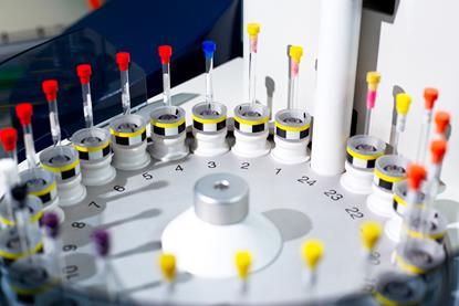 Close-up of a laboratory sample analysis machine with multiple vials in a circular arrangement, featuring color-coded caps in red, yellow, and blue. The equipment is used for automated testing or sample processing in scientific or medical research