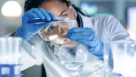 Close up of a young female scientist pouring a blue chemical from a vial into a round bottom flask